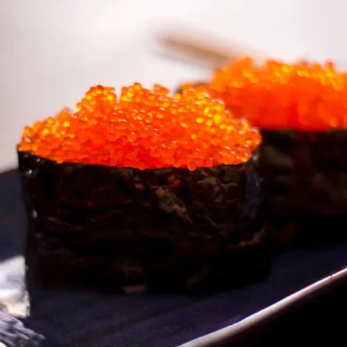 Fresh ingredients for Tobiko Sushi, including sushi-grade fish, nori sheets, tobiko eggs, cucumber, and sushi rice arranged on a wooden cutting board